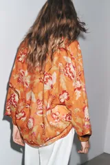 Camisa arrugada de ramio color naranja con estampado floral en tonos rosados y blancos. Tiene cuello redondo con escote pico ajustable con lazadas, mangas 3/4 y bajo asimétrico con aberturas laterales.