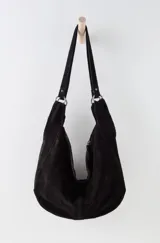 Bolso tipo hobo de gamuza negra, con forma holgada y cierre principal de cremallera. Se lleva al hombro con dos asas de correa delgadas sujetas con argollas metálicas plateadas.