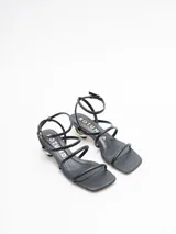 Sandalias negras de cuero con tiras finas, taco geométrico dorado y pulsera al tobillo.