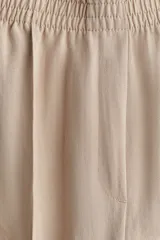Pantalón de corte holgado color beige, confeccionado en sarga de mezcla de lyocell. Modelo de tiro alto con cintura fruncida, cierre decorativo y bolsillos laterales discretos. Piernas rectas con pliegues cosidos adelante y raya planchada atrás.