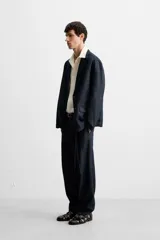Pantalón de denim azul oscuro, corte wide fit, largo cropped, con pliegues frontales, bolsillos laterales y traseros asimétricos. Cierre frontal con cremallera y gancho metálico oculto.