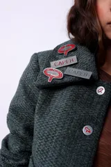 Campera corta de poliéster texturizado color verde oscuro, con cuello camisero, cierre frontal con botones a presión, bolsillos delanteros y forro interior. Incluye pins decorativos.