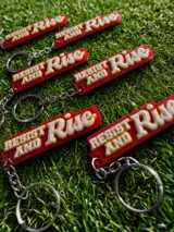 Llavero rectangular con cadena y aro metálico. Diseño con la frase "Resist and Rise" en letras blancas con borde amarillo sobre fondo rojo.