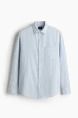 Camisa de hombre de corte holgado, confeccionada en tela de sirsaca a rayas verticales celestes y blancas. Tiene cuello inglés, tapeta clásica con botones, bolsillo de parche en el pecho, mangas largas con puños abotonados y bajo redondeado.