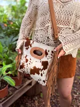 Bandolera de cuero con estampado animal print de vaca en tonos marrón y blanco, con flecos de cuero marrón en los laterales. Tiene correa larga de cuero marrón regulable y asa de mano con forma ovalada.