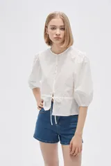 Blusa blanca de algodón con textura de lunares, corte recto, cuello redondo, mangas 3/4 abullonadas y cierre frontal con botones.