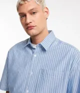 Camisa masculina oversized boxy de manga corta, confeccionada en tejido oxford a rayas verticales celestes y blancas. Cuenta con cuello inglés, abotonadura frontal y bolsillo de parche en el pecho.