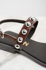 Sandalias bajas de cuero color marrón con apliques de tachas metálicas plateadas.