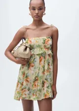 Vestido corto de gasa color amarillo pálido con estampado de rosas anaranjadas y hojas verdes. Tiene escote recto con volantes, tirantes finos y cierre lateral con cremallera.