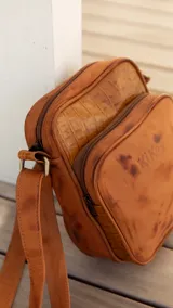 Bandolera de cuero color terracota con correa ajustable, cierre superior y bolsillo externo con cierre. Incluye bolsillo interno y logo de la marca en el frente.