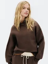 Buzo con capucha de color marrón oscuro, de corte holgado y mangas largas con costuras visibles. Se vislumbra un borde naranja debajo del buzo y un pantalón de jogging marrón con cordón blanco en la cintura.