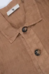 Campera color marrón claro estilo sobrecamisa confeccionada en lino premium. Cuenta con cuello clásico, cierre frontal con botones, un bolsillo de parche en el pecho y dos bolsillos de parche en la parte inferior.