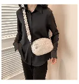 Cartera bandolera pequeña color beige, de cuero sintético con textura granulada. Tiene correa ancha de tela estampada con motivos geométricos y cierre superior con cremallera. Incluye un bolsillo frontal con cremallera y un dije metálico.