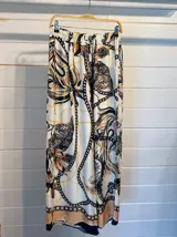 Pantalón ancho de satén con cintura elástica y estampado tipo pañuelo con motivos de cadenas y hojas en tonos blanco, negro, marrón y dorado. Presenta un borde inferior en color beige/durazno.