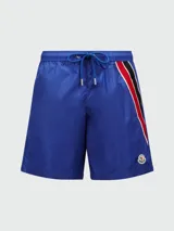 Short de baño azul con cintura elástica y cordón ajustable. Presenta una franja tricolor (rojo, negro y blanco) en el lateral y el logo de la marca en el ruedo.