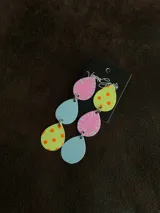 Aros colgantes con tres figuras geométricas: gota fucsia con detalles dorados, círculo dorado y gota verde con lunares fucsias.