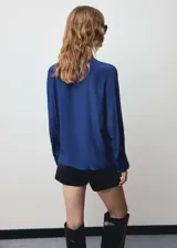 Blusa azul con lazo al cuello, de tejido fluido y satinado, con cierre oculto de botones y mangas largas abotonadas.