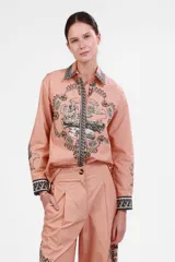 Camisa de popelina rosa con estampado de paisajes y figuras en blanco y negro. Tiene cuello clásico, cierre frontal con botones y mangas largas con puños abotonados.