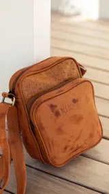 Bandolera de cuero color terracota con correa ajustable, cierre superior y bolsillo externo con cierre. Incluye bolsillo interno y logo de la marca en el frente.