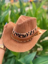 Sombrero de ala ancha tejido en hilo de coco y sintético, color marrón claro. La copa está cubierta con encaje naranja quemado con patrón floral. Tiene una cinta de cuero marrón oscuro y un aplique decorativo de hoja de gamuza con incrustaciones de pedrería.