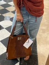 Bolso tipo tote bag de gamuza color marrón óxido, con panel central de cuero con textura de cocodrilo en tono marrón oscuro. Posee asas superiores de cuero marrón oscuro y una correa de hombro desmontable a juego. El cierre principal es metálico tipo pestillo.