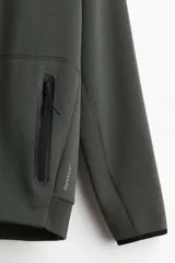 Campera bomber deportiva de tejido funcional color verde oscuro, con cuello elevado corto, cierre bidireccional y mangas raglán. Posee bolsillos delanteros con cierre y costuras selladas.