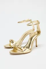 Sandalias doradas de tacón fino con tira delantera con detalle de lazo y tira con hebilla al tobillo.