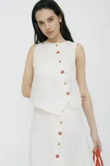 Top sin mangas color crudo, confeccionado en lino 100%, con cartera asimétrica y botones de madera. Tiene ojales bordados con forma de frutillas.