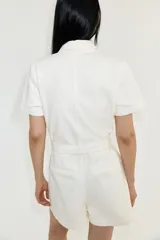 Mono corto color blanco de denim rígido, con cuello camisero, cierre abotonado, mangas cortas, bolsillos superiores con botón y cinturón extraíble.