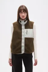 Chaleco sin mangas de textura corderito (fleece) color marrón oscuro, con cuello alto y cierre frontal de botones a presión. Presenta un bolsillo superior con cierre en el lado izquierdo del pecho, con solapa de tela lisa de color verde claro.