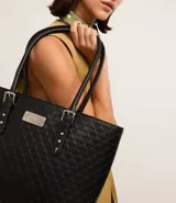 Bolso shopper grande de poliuretano negro con textura acolchada. Tiene correa de mano con detalles de hebilla y remaches plateados. Cierre superior con cremallera y forro de poliéster.