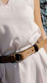 Cinturón de cuero de dos tonos, con una sección principal de cuero color camel claro con perforaciones y una sección más oscura (marrón oscuro) que pasa por la hebilla. La hebilla es metálica, redonda y de color oscuro.