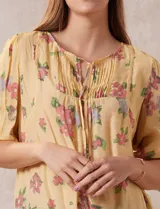 Blusa amarilla estampada con flores rosadas, de gasa, con cuello a la base, escote en V plisado y lazo de ajuste. Delantera con detalles en puntilla y mangas 3/4 con puño fruncido.