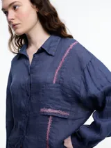 Camisa de manga larga de lino color azul con calce relajado y diseño asimétrico. El bolsillo delantero está colocado de forma invertida, un detalle lúdico que rompe con lo clásico. Lleva un bordado tipo pespunte en color contraste que recorre el frente y el bolsillo.