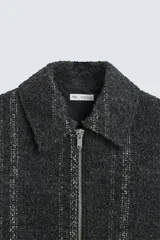 Sobrecamisa negra con textura de tejido bouclé y rayas verticales plateadas. Tiene cuello solapa, manga larga con puños abotonados, bolsillos laterales y cierre frontal con cremallera.