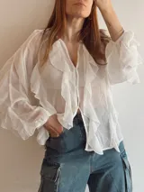 Blusa blanca traslúcida con cuello en V, cierre frontal con botones y mangas largas con volados.