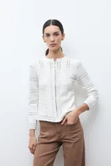 Chaqueta de tejido punto color blanco crudo, de manga larga y cuello redondo, con cierre frontal metálico. Presenta un diseño calado con aberturas verticales a lo largo del cuerpo y las mangas.
