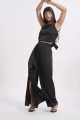 Pantalón de vestir color beige claro, tiro alto, de corte ancho y pierna recta. Presenta un detalle de tela drapeada o solapa larga que cae desde la cintura sobre la pierna izquierda.