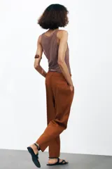 Pantalón color marrón confeccionado en hilatura con mezcla de lyocell y algodón. Tiro medio, cintura con trabillas, bolsillos laterales, pinzas en delantero y raya planchada. Cierre frontal con cremallera, botón y ganchos metálicos.