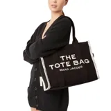 Bolso tote grande de tela jacquard color negro con estampado del logo "THE TOTE BAG MARC JACOBS" en blanco. Tiene doble asa de mano, laterales con logo repetido y cierre superior con cremallera.