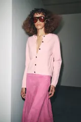 Chaqueta de punto color rosa pálido, con cuello redondo y cierre frontal de botones oscuros. Presenta mangas largas y un corte a la cintura.