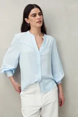 Camisa celeste de manga larga con cuello redondo y escote en V. Presenta detalles de pasamanería en el frente y las mangas, y botones forrados al tono. De corte fluido.