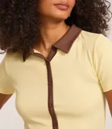 Blusa color amarillo claro confeccionada en tejido rib, con cuello polo marrón, manga corta y cierre frontal con botones marrones.