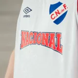 Camiseta de básquet Umbro del Club Nacional de Football, modelo oficial. Diseño blanco con detalles en azul y rojo en cuello y sisas. Incluye logo de Umbro, escudo del club y sponsor Antel en el frente.