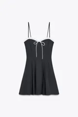 Vestido corto negro con tirantes finos ajustables, escote recto con volantes blancos y lazo decorativo. Cierre lateral con cremallera oculta.