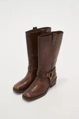 Bota de caña media color negro, estilo motociclista o cowboy, con puntera cuadrada y taco bajo. Presenta una tira ancha con hebillas metálicas alrededor del tobillo.