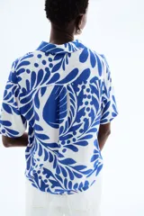 Camisa de manga corta con estampado de hojas y flores en tonos azules sobre fondo blanco. Tiene cuello clásico, botones frontales, corte holgado y canesú con pliegue en la espalda.
