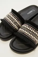 Sandalias tipo slider blancas, con tira ancha decorada con trenzado de hilo beige y plateado, tachas piramidales plateadas y flecos blancos en los bordes.