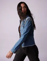 Campera de jean celeste de la marca Baccini, con cierre central, bolsillos delanteros, apliques colgantes de tachas en los hombros y terminación desflecada en el ruedo.