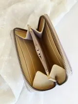 Billetera pequeña de cuero color crema, con cierre metálico y monedero interno. Presenta un diseño de cruces marrones en el frente y textura símil cocodrilo en un lateral.